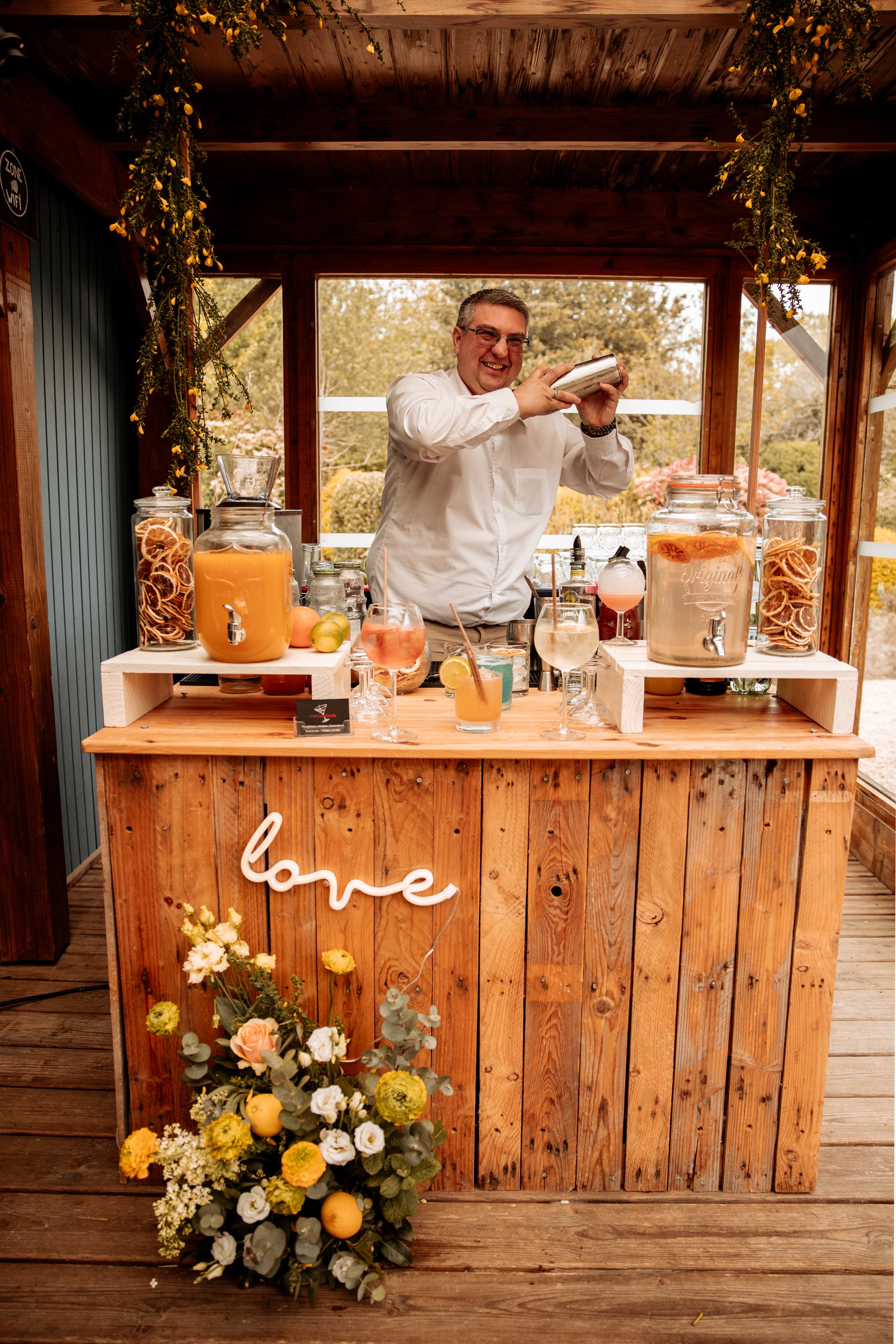 Bar à cocktails en bois décoré de fleurs jaunes et d'agrumes, avec un barman au shaker, lors d'un mariage sur le thème Dolce Vita